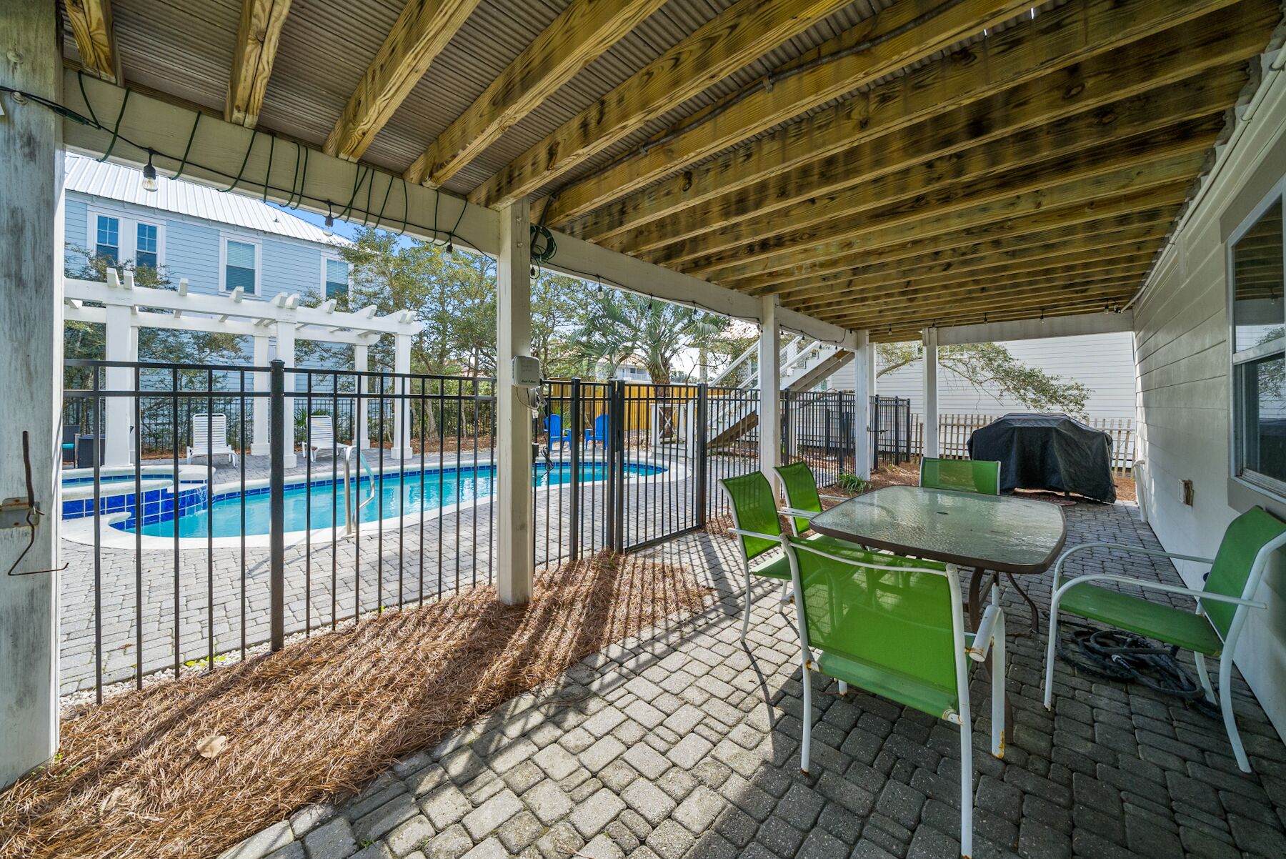 A covered patio with a pool, green chairs, and a barbecue grill.