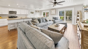 A modern kitchen with white cabinets, a microwave, and a stove. There is a large gray sectional sofa with pillows and a wooden coffee table. The room has wooden floors and a ceiling fan. - Joanna House! Seagrove Beach Cottage, Private Pool, Spacious, close to Seaside. (Seagrove Beach)