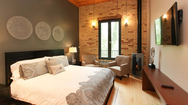 Un dormitorio con una cama, dos sillones, una mesa, chimenea y un ventanal grande.