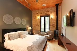 Un dormitorio con una cama, dos sillones, una mesa, chimenea y un ventanal grande.
