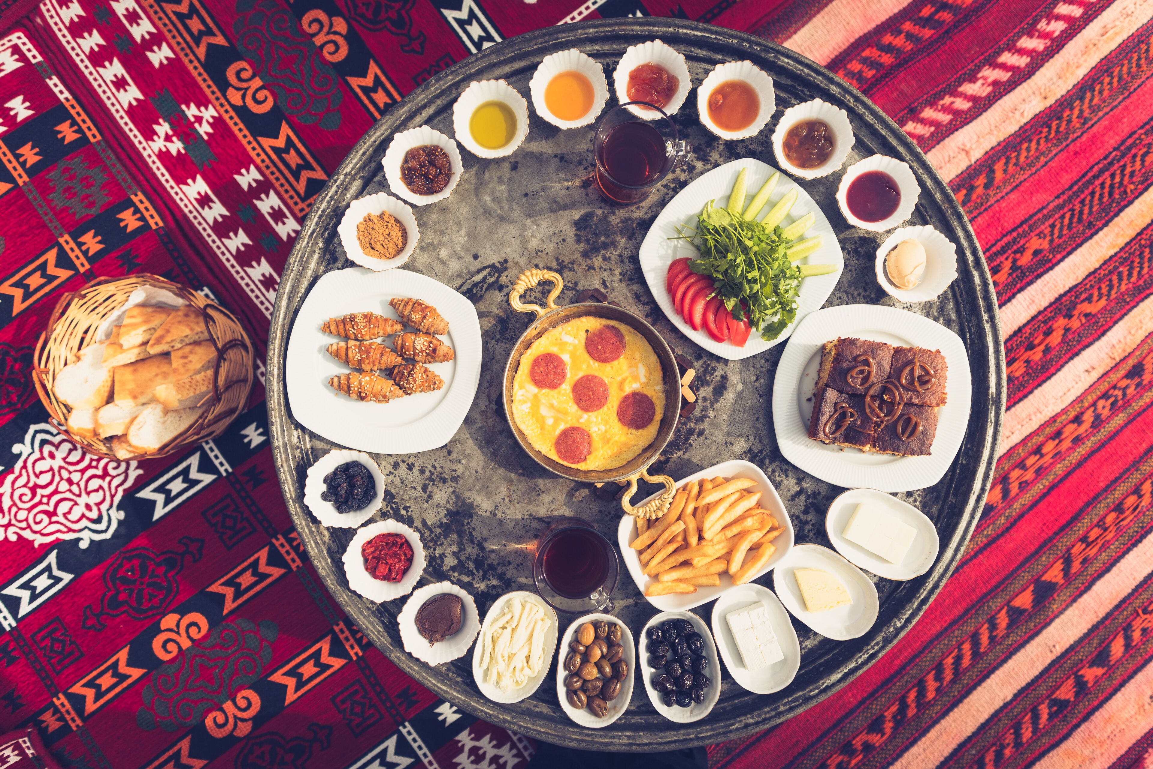 A round tray with various food items and condiments, including bread, pastries, and a variety of dips, arranged on a patterned tablecloth.