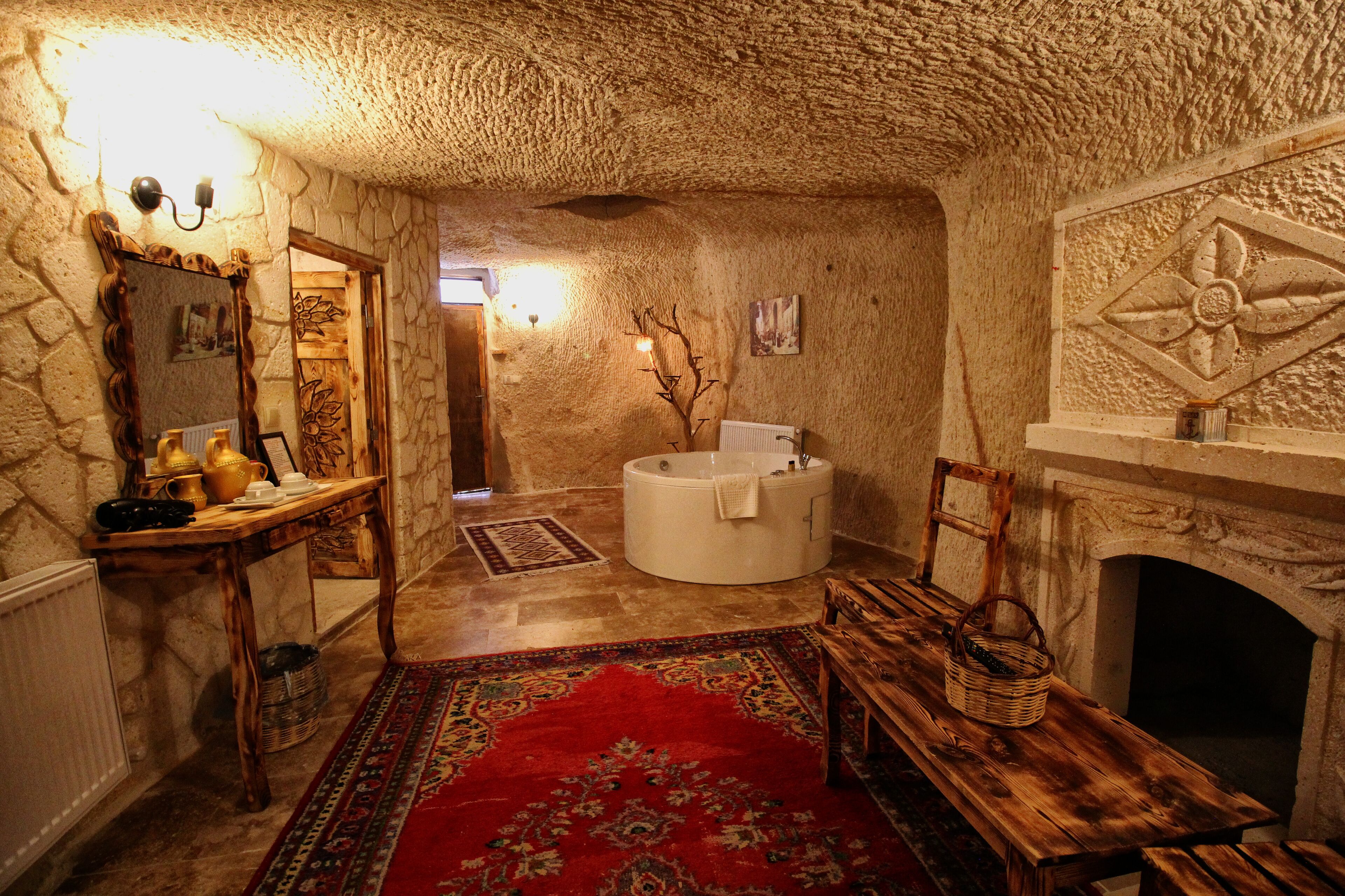 Una habitación rústica con paredes de piedra, un banco de madera, un tapete rojo estampado, una chimenea de piedra y una bañera redonda.