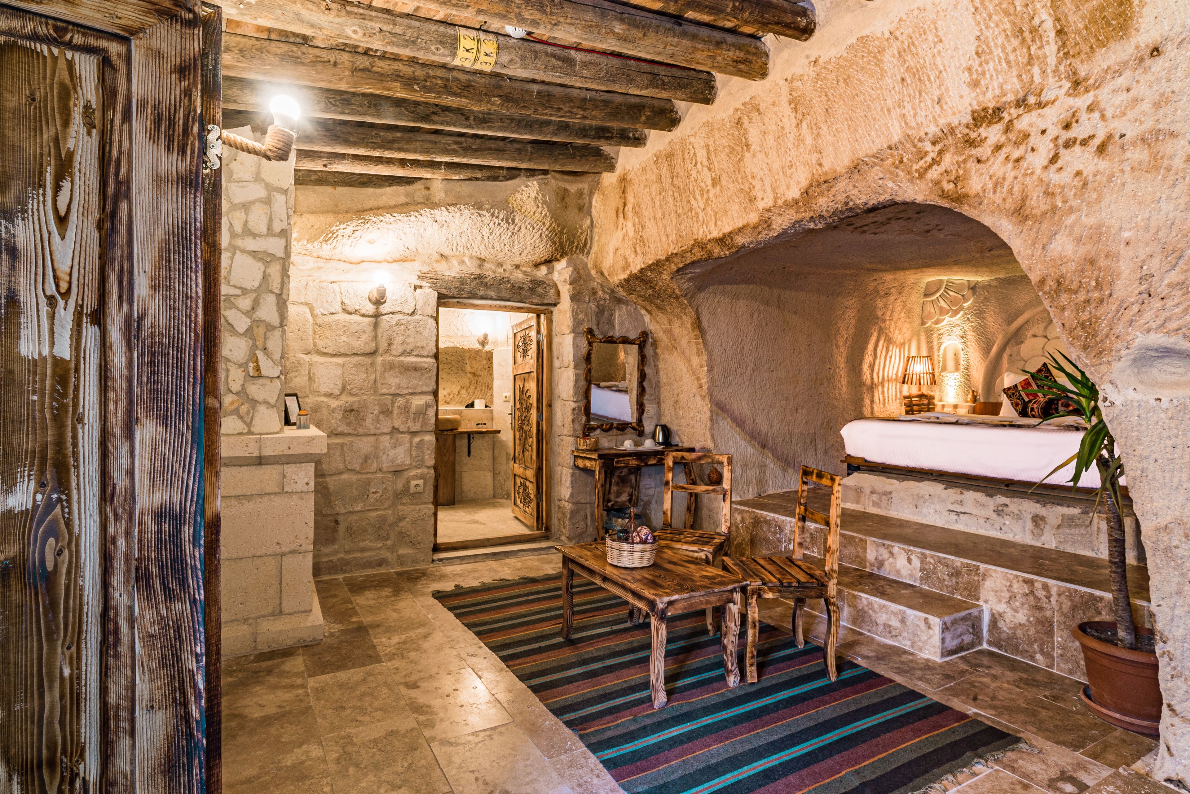 A rustic stone interior with a wooden bench, a striped rug, and a stone fireplace.