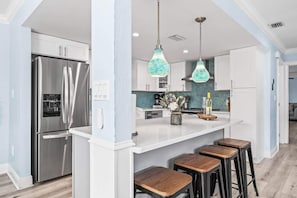 A modern kitchen with a stainless steel refrigerator, white cabinets, a marble countertop, and wooden bar stools.