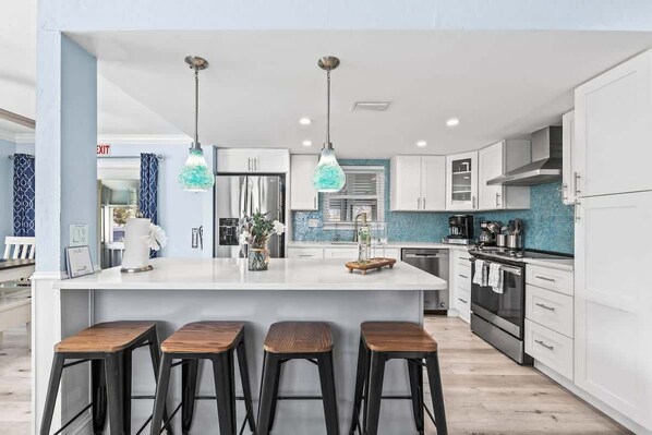 A modern kitchen with a central island, bar stools, stainless steel appliances, and blue-tiled backsplash.