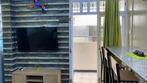 Un salon doté d’un téléviseur à écran plat fixé sur un mur imitant la pierre, d’un meuble de rangement en bois, d’un coin repas avec une table en granit et d’une porte-fenêtre coulissante donnant sur un balcon.