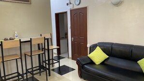 Un espace de vie comprenant un canapé en cuir noir, une table à manger en bois avec des chaises, un coin cuisine avec une horloge, et une porte donnant sur une autre pièce.
