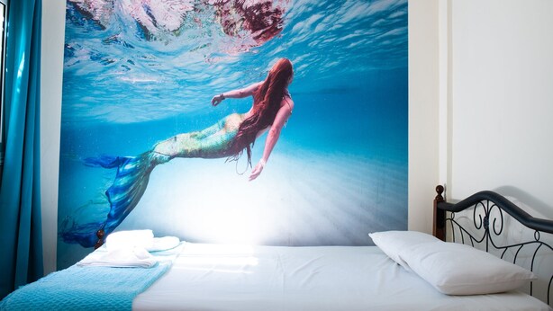 Een slaapkamer met een muurschildering van een zeemeermin, een bed met witte lakens en een raam met blauwe gordijnen.