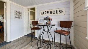 Tunnelmallinen ruokailutila, jossa on koristeena kyltti, jossa lukee ”Farmhouse”, kaksi metallituolia, pieni pöytä, jossa on maljakko ja kynttilä, sekä kyltti, jossa lukee: ”Kootkaa yhteen iloisin mielin”.