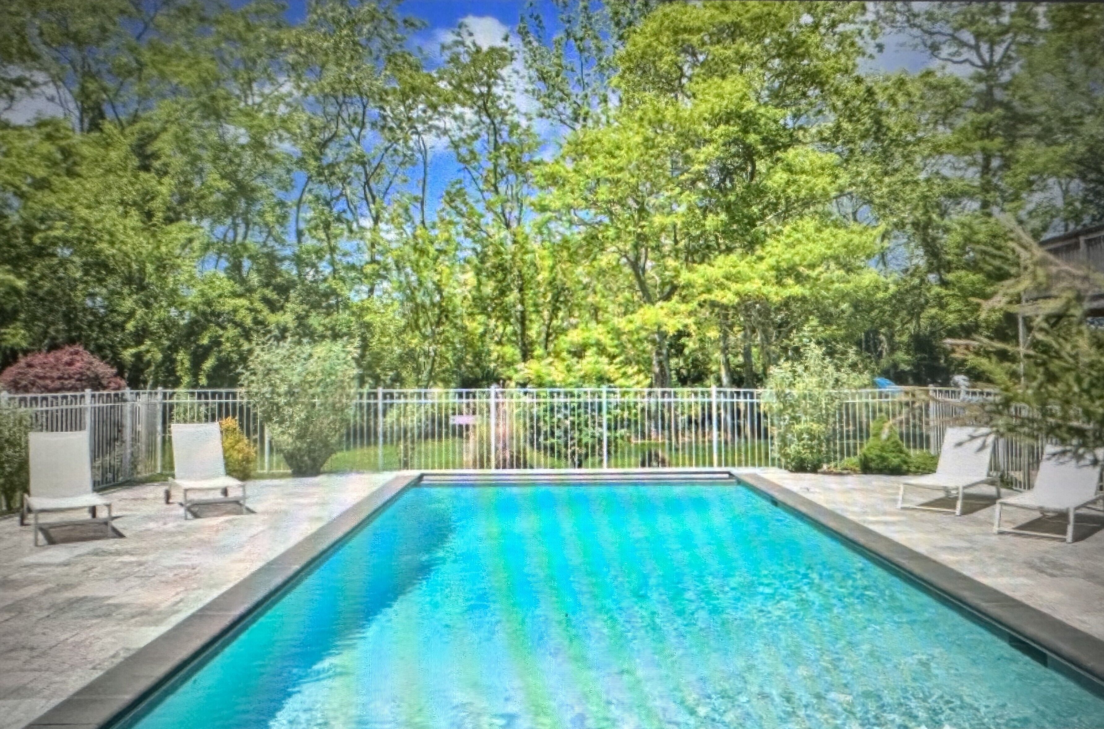 Une piscine rectangulaire à l’eau d’un bleu cristallin, entourée d’une clôture blanche et de chaises longues blanches.