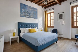 Une chambre avec un lit bleu, des oreillers blancs et jaunes, un tableau au mur et une fenêtre avec des rideaux.
