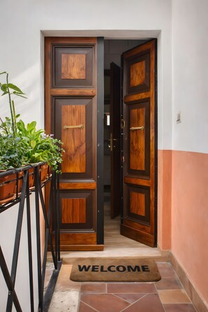 A wooden door with a 'WELCOME' mat, a potted plant, and a tiled floor. - Two Bedroom Apartment (Roma)