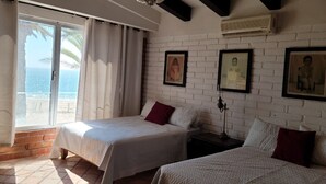Una habitación con dos camas, un ventanal con cortinas, una pared de ladrillo y cuadros enmarcados.