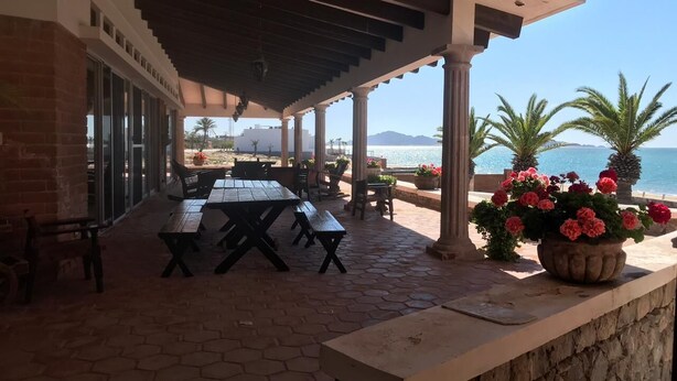 Un patio al aire libre con mesas y sillas, un muro de piedra y vista al mar.