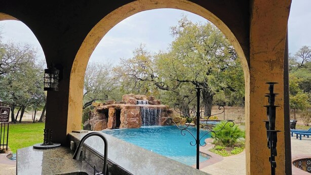 An outdoor pool area with a waterfall feature, surrounded by arches and greenery.