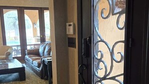A foyer with a dark door featuring ornate metalwork, a patterned rug, and a view into a living area with a sofa and a ceiling fan.
