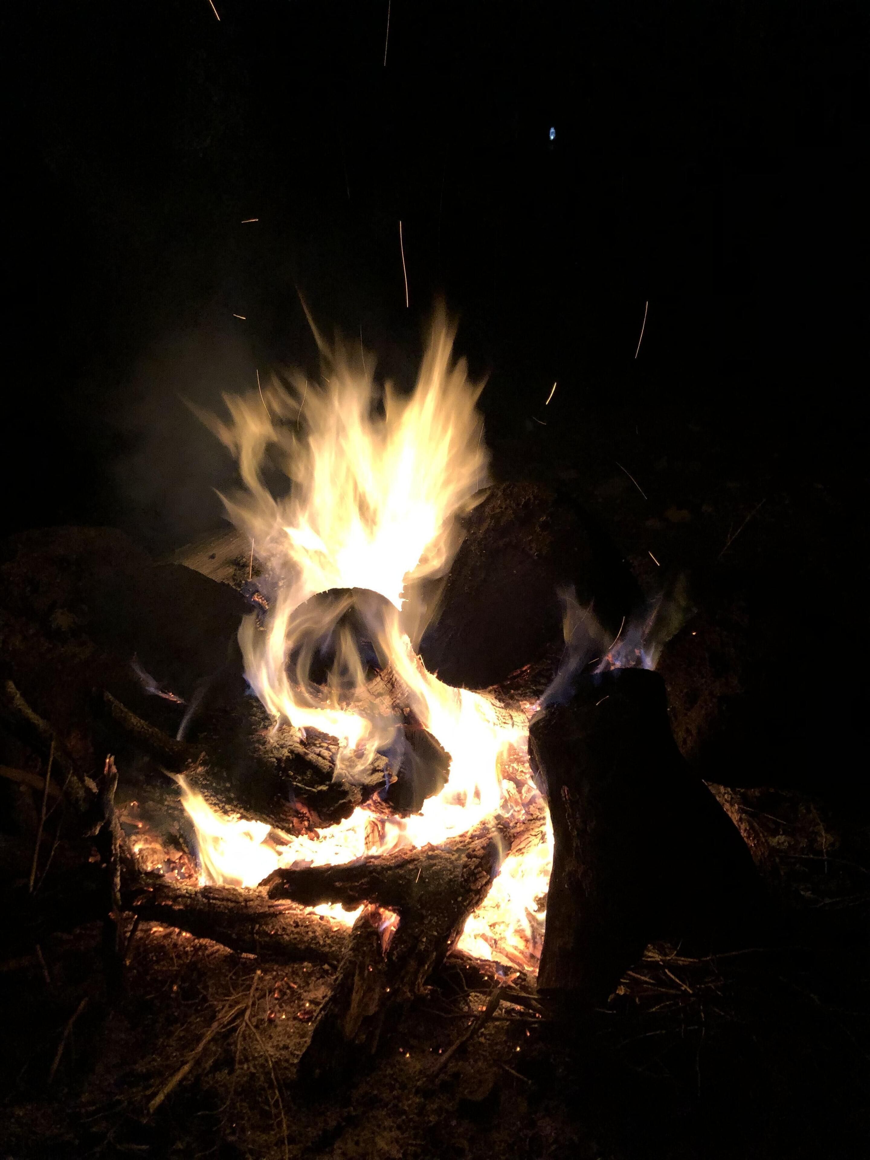 Uma fogueira com lenha e brasas, rodeada por chão e pedras naturais.