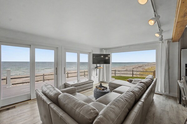 A modern living room with a large sofa, a flat-screen TV, and a view of the beach.