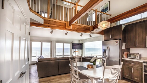 A spacious kitchen with a wooden staircase, a dining area with white chairs, and a view of the ocean through large windows. - 14 Captain Crocker (East Sandwich)