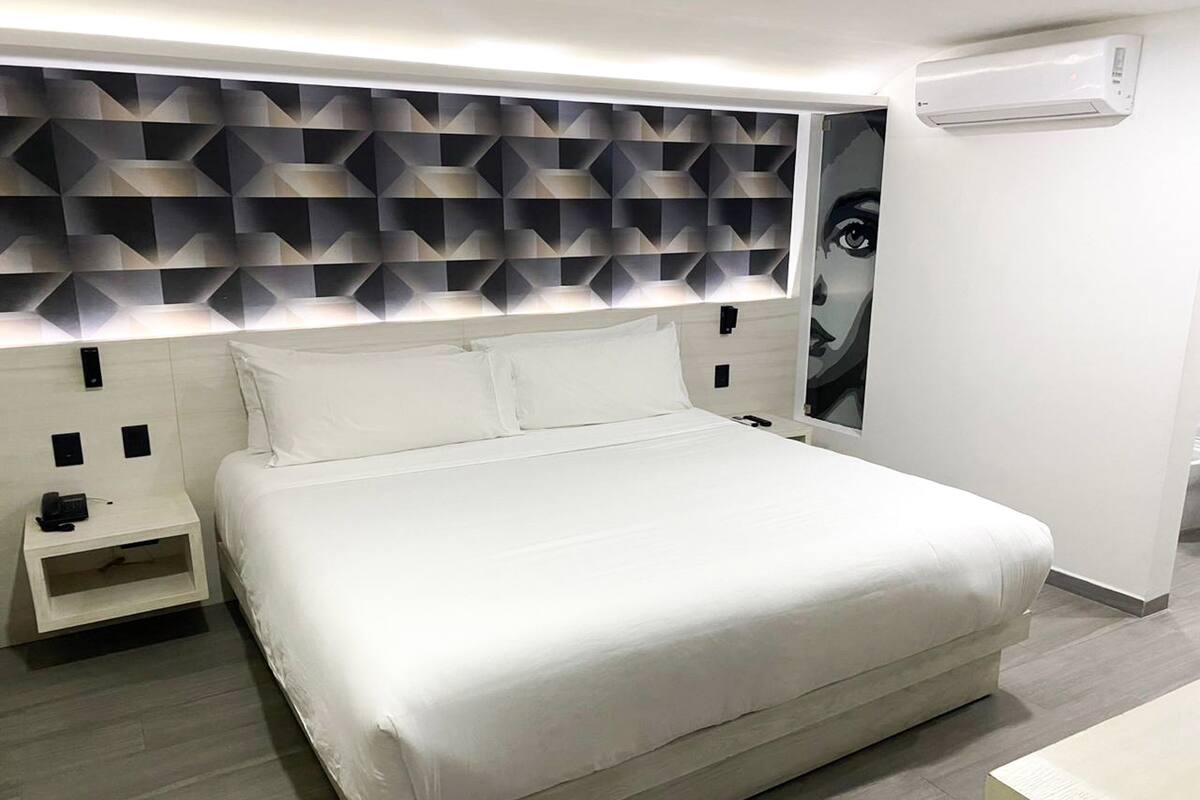 A modern hotel room with a large bed, a bedside table, a wall-mounted air conditioner, and a geometric-patterned headboard.