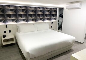 Una habitación de hotel moderna con una cama grande, una mesita de noche, aire acondicionado de pared y un cabecero con motivos geométricos.