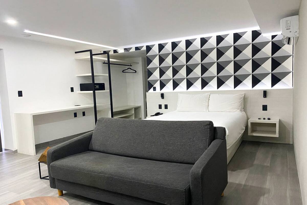 A modern hotel room with a grey sofa, a bed with white bedding, a geometric-patterned wall, and a small bedside table.