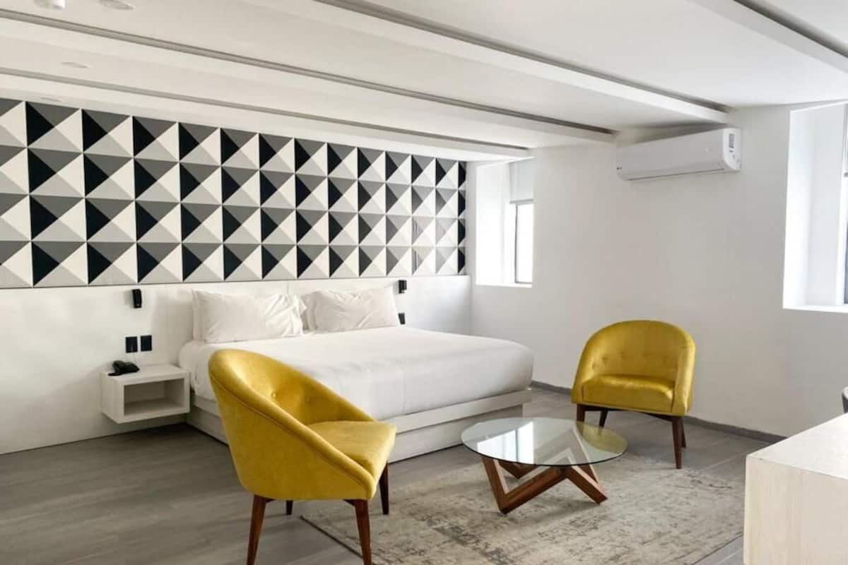 A modern hotel room with a white bed, two yellow armchairs, a glass coffee table, and a geometric-patterned wall.