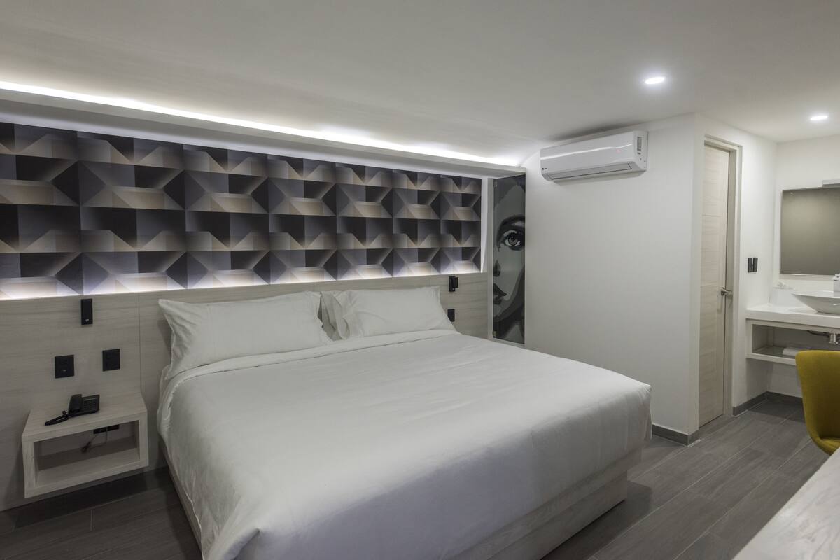 A modern hotel room with a large bed, a bedside table with a phone, a wall-mounted air conditioner, and a bathroom with a mirror and sink.