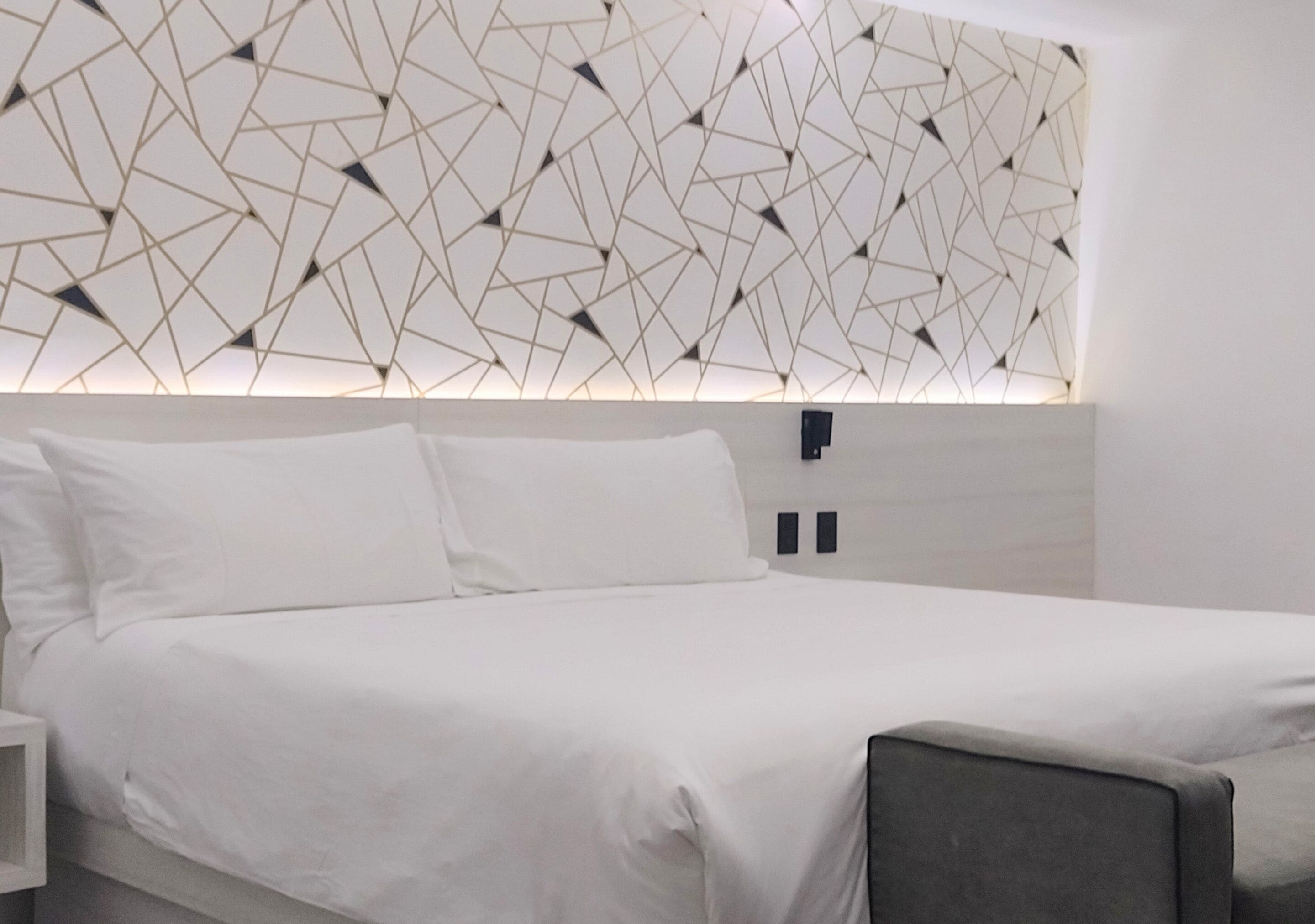 Quarto de hotel moderno com parede de padrão geométrico, uma cama bem arrumada e uma pequena mesa de cabeceira.
