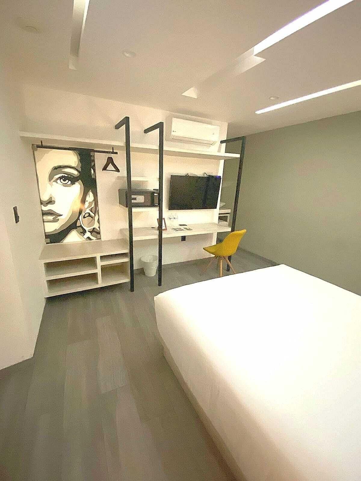 A modern bedroom with a large bed, a TV mounted on the wall, and a unique artwork on the left side.