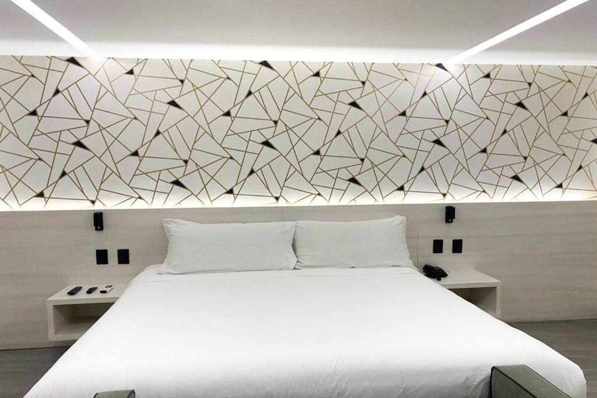 A modern hotel room with a geometric-patterned headboard, a large bed with white linens, and a bedside table with a phone and remote controls.