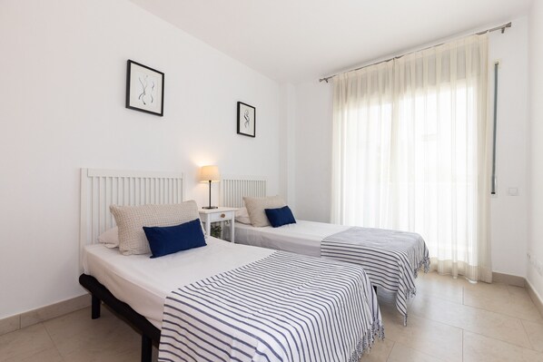 Habitación de hotel con dos camas, una mesita de noche con lámpara y un ventanal con cortinas translúcidas.
