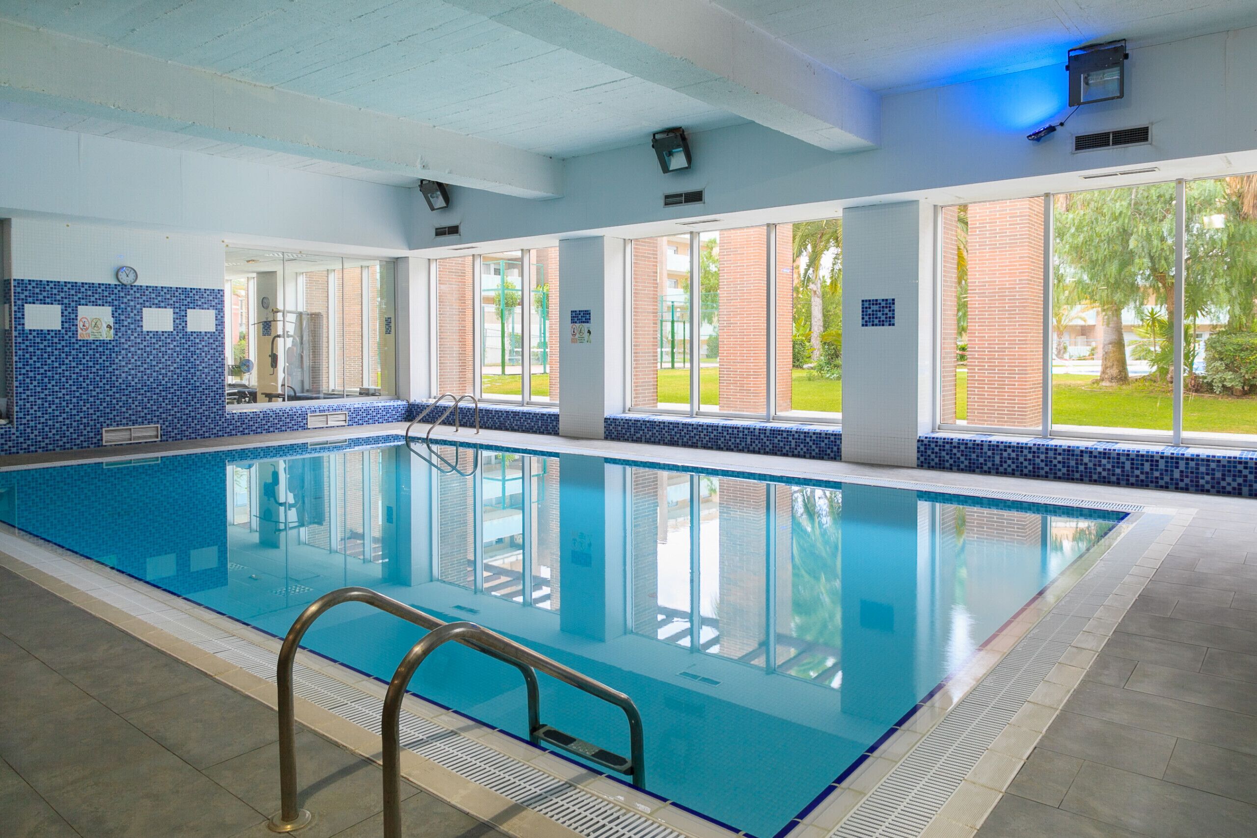 An indoor swimming pool with clear glass walls, a tiled floor, and a metal ladder.