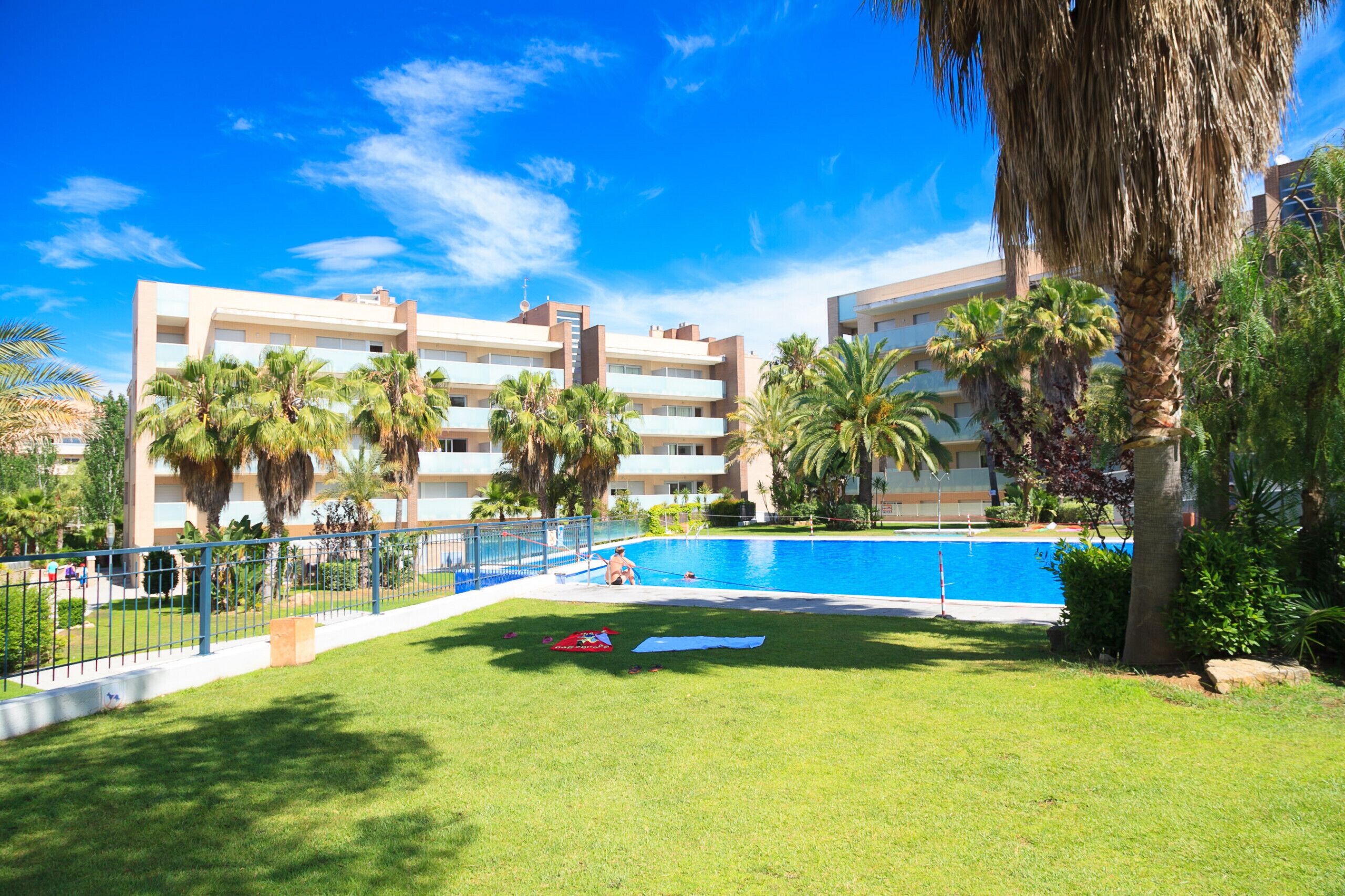 A hotel with a swimming pool, palm trees, and a clear blue sky.