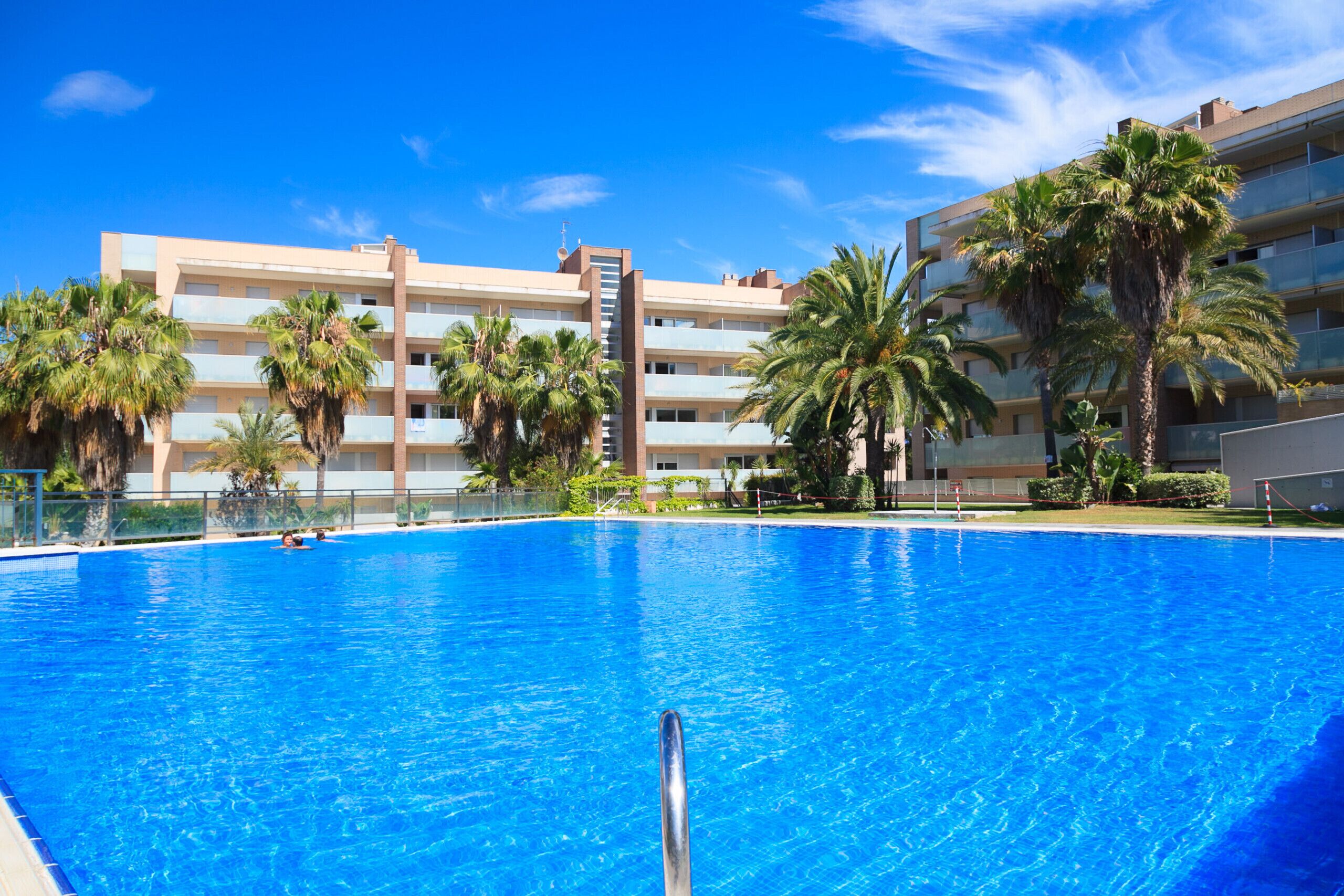 A swimming pool with a clear blue water surface, surrounded by palm trees and a multi-story building with balconies.