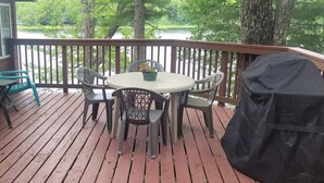 A wooden deck with a round table and chairs, a covered grill, and a view of a lake. - Beautiful Lakefront Cottage with Mountain Views (Dedham)