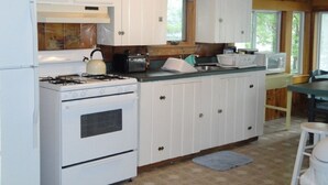 A compact kitchen with white cabinets, a stove, a sink, and a window. - Beautiful Lakefront Cottage with Mountain Views (Dedham)