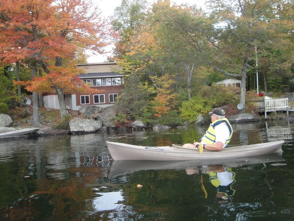 A person in a canoe on a calm body of water, surrounded by autumn-colored trees and a building with a fence. - Beautiful Lakefront Cottage with Mountain Views (Dedham)