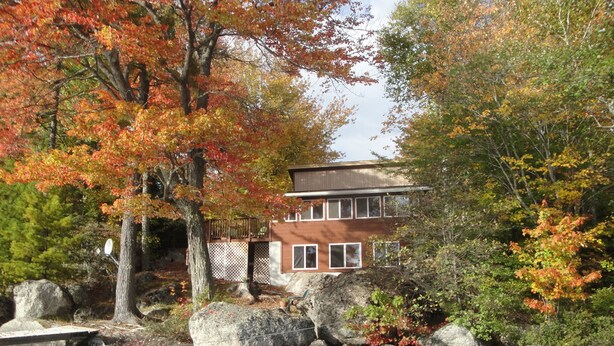 A house with a deck, surrounded by autumn foliage and a pond. - Beautiful Lakefront Cottage with Mountain Views (Dedham)