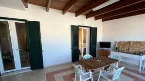 Un espace patio avec une table en bois et des chaises blanches, une porte-fenêtre coulissante et un canapé blanc doté d’une tête de lit en bois.