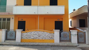 Un bâtiment à deux étages avec une façade jaune, une entrée surmontée d’un mur en pierre et une allée menant à une porte grillagée.