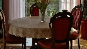 A dining area with a table set for two, a vase with flowers, and a view of a green landscape through large windows.