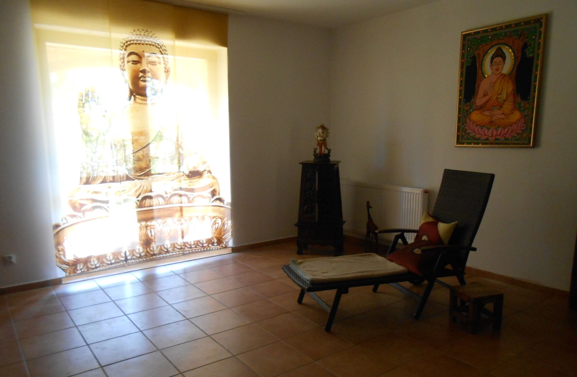 A room with a large Buddha statue, a chair, a small table, and a framed picture on the wall.