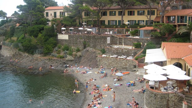 A beach with people sunbathing and swimming, a restaurant named 'RESTAURANT LA BALETTE', and buildings with red-tiled roofs.