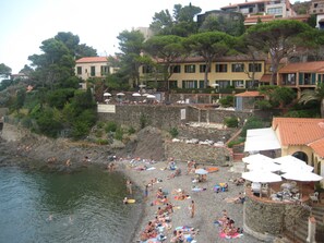 A beach with people sunbathing and swimming, a restaurant named 'RESTAURANT LA BALETTE', and buildings with red-tiled roofs.