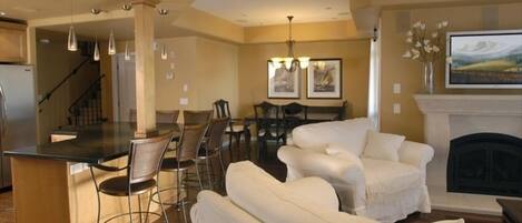 A modern living room with a fireplace, a dining area with a chandelier, and a kitchen with a bar.