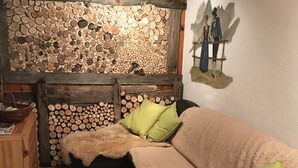 A cozy room with a wooden ceiling, a wall decorated with stacked logs, a sofa with fluffy cushions, and a painting of two people on a bench.