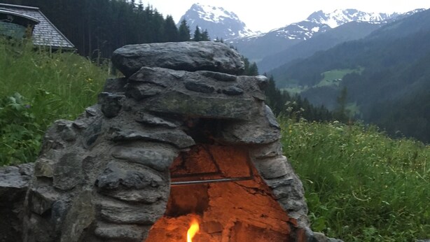 A stone-built outdoor oven with a fire burning inside, surrounded by a stone wall and set against a mountainous backdrop.