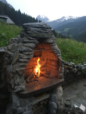 A stone-built outdoor oven with a fire burning inside, surrounded by a stone wall and set against a mountainous backdrop.