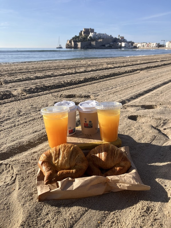 A beach scene with a tray of croissants and orange juice cups on the sand, with a distant view of a coastal town.